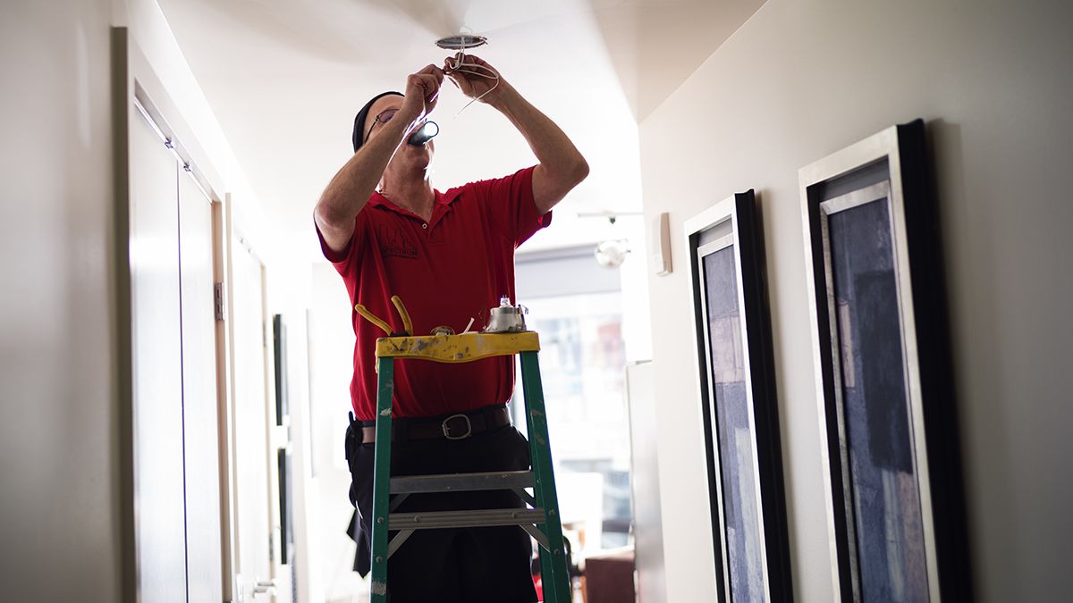 handyman standing on ladder fixing light fixture on ceiling in a hallway