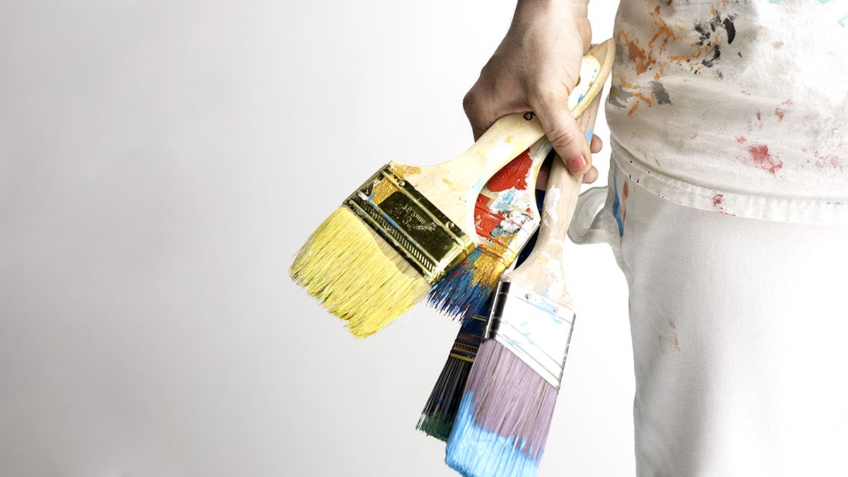 person with paint splatter on shirt holding multiple paint brushes of different sizes and colors